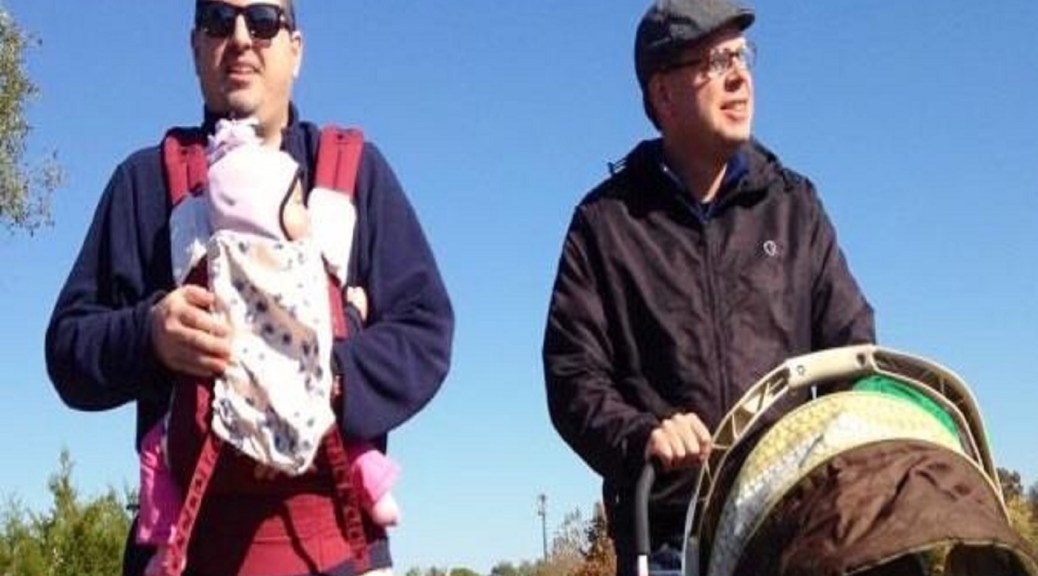 Two 30-something men stand side-by-side, Evan with his daughter in a sling, and Tony with his son in a stroller.